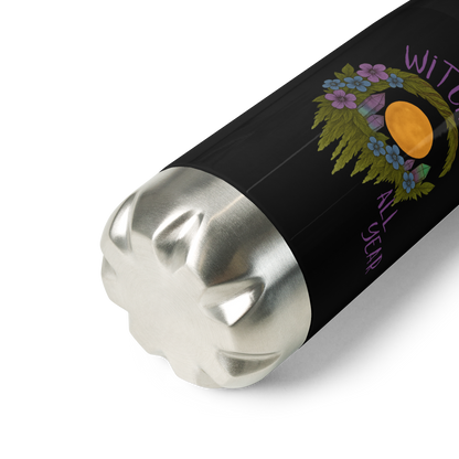 This is a zoom in of a black stainless steel water bottle  with a design of a hand-drawn wreath made of moss-covered branches wraps around a glowing sun. The wreath is decorated with purple and blue flowers, small amethyst and fluorite crystals, and strands of hanging moss. The words “Witchy” curve above the wreath and “All Year” sits on the bottom right 