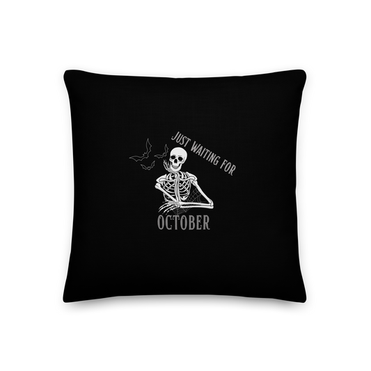 This is a black throw pillow that has a bored skeleton with his head resting in his hand and he has spider webs and bats around him and the text reads just waiting for October