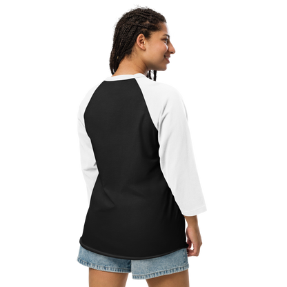 This is a woman with dark braids wearing a black and white raglan shirt with a Día de los Muertos–style illustration of a male and female skeleton holding hands, surrounded by marigolds, vines, and folk art patterns. The skeletons wear traditional black clothing, the man in a suit and hat, the woman with a floral crown with a red heart and leaves at the bottom and the text Creepy All Year. Rendered in warm tones of orange, green, and cream, symbolizing eternal love and remembrance on the front side