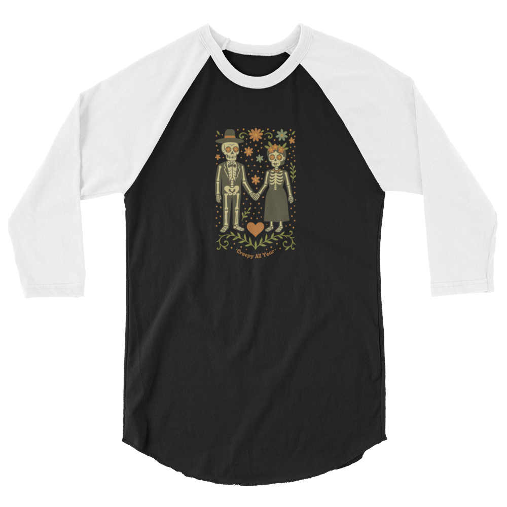 This is a black and white raglan shirt with a Día de los Muertos–style illustration of a male and female skeleton holding hands, surrounded by marigolds, vines, and folk art patterns. The skeletons wear traditional black clothing, the man in a suit and hat, the woman with a floral crown with a red heart and leaves at the bottom and the text Creepy All Year. Rendered in warm tones of orange, green, and cream, symbolizing eternal love and remembrance