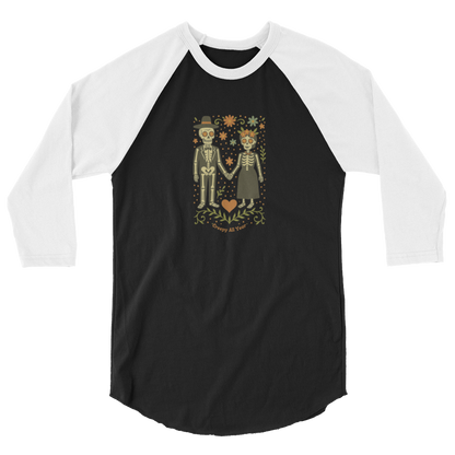This is a black and white raglan shirt with a Día de los Muertos–style illustration of a male and female skeleton holding hands, surrounded by marigolds, vines, and folk art patterns. The skeletons wear traditional black clothing, the man in a suit and hat, the woman with a floral crown with a red heart and leaves at the bottom and the text Creepy All Year. Rendered in warm tones of orange, green, and cream, symbolizing eternal love and remembrance