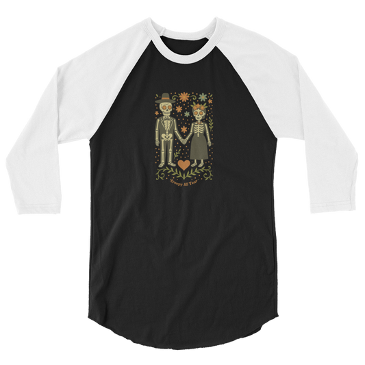 This is a black and white raglan shirt with a Día de los Muertos–style illustration of a male and female skeleton holding hands, surrounded by marigolds, vines, and folk art patterns. The skeletons wear traditional black clothing, the man in a suit and hat, the woman with a floral crown with a red heart and leaves at the bottom and the text Creepy All Year. Rendered in warm tones of orange, green, and cream, symbolizing eternal love and remembrance
