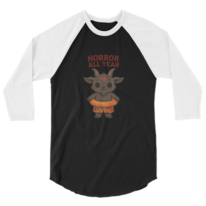 This is a black and white raglan with a cartoon illustration of a baby Baphomet goat with curved horns and a playful grin, wearing bright floral swim trunks while floating in a colorful inner tube. The character is set against a light background with the bold text “Horror All Year” above in red letters