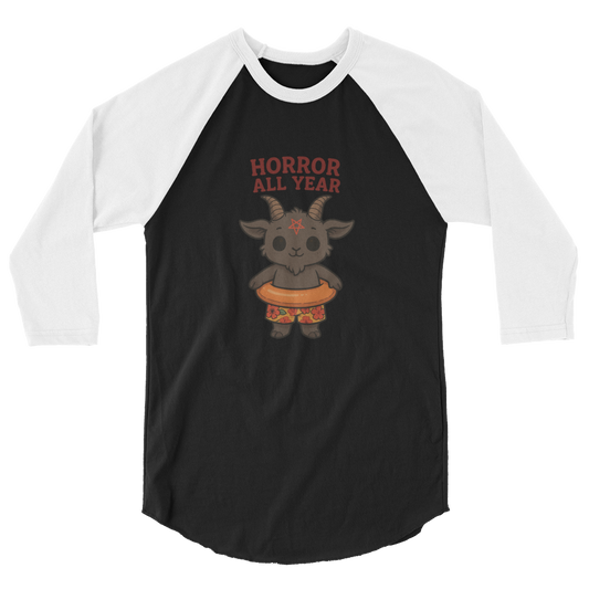 This is a black and white raglan with a cartoon illustration of a baby Baphomet goat with curved horns and a playful grin, wearing bright floral swim trunks while floating in a colorful inner tube. The character is set against a light background with the bold text “Horror All Year” above in red letters