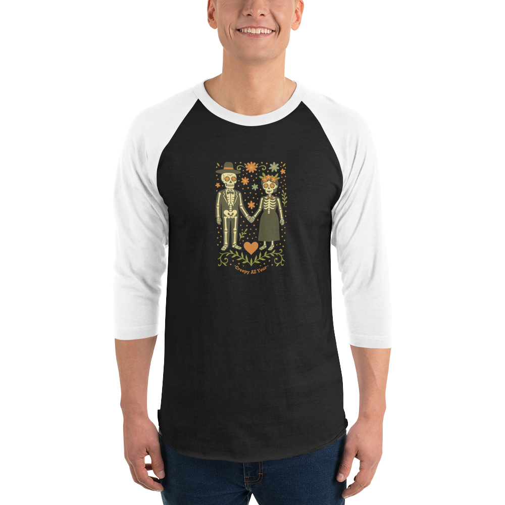 This is a man wearing a black and white raglan shirt with a Día de los Muertos–style illustration of a male and female skeleton holding hands, surrounded by marigolds, vines, and folk art patterns. The skeletons wear traditional black clothing, the man in a suit and hat, the woman with a floral crown with a red heart and leaves at the bottom and the text Creepy All Year. Rendered in warm tones of orange, green, and cream, symbolizing eternal love and remembrance