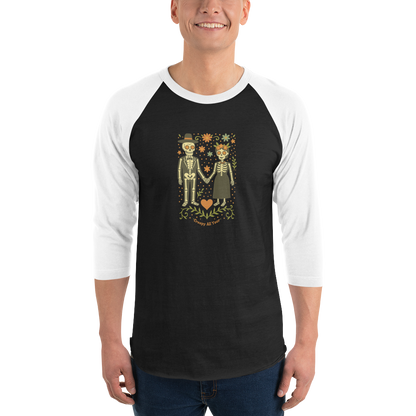 This is a man wearing a black and white raglan shirt with a Día de los Muertos–style illustration of a male and female skeleton holding hands, surrounded by marigolds, vines, and folk art patterns. The skeletons wear traditional black clothing, the man in a suit and hat, the woman with a floral crown with a red heart and leaves at the bottom and the text Creepy All Year. Rendered in warm tones of orange, green, and cream, symbolizing eternal love and remembrance