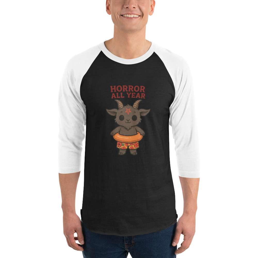 This is a man wearing a black and white raglan with a cartoon illustration of a baby Baphomet goat with curved horns and a playful grin, wearing bright floral swim trunks while floating in a colorful inner tube. The character is set against a light background with the bold text “Horror All Year” above in red letters