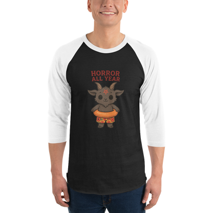 This is a man wearing a black and white raglan with a cartoon illustration of a baby Baphomet goat with curved horns and a playful grin, wearing bright floral swim trunks while floating in a colorful inner tube. The character is set against a light background with the bold text “Horror All Year” above in red letters