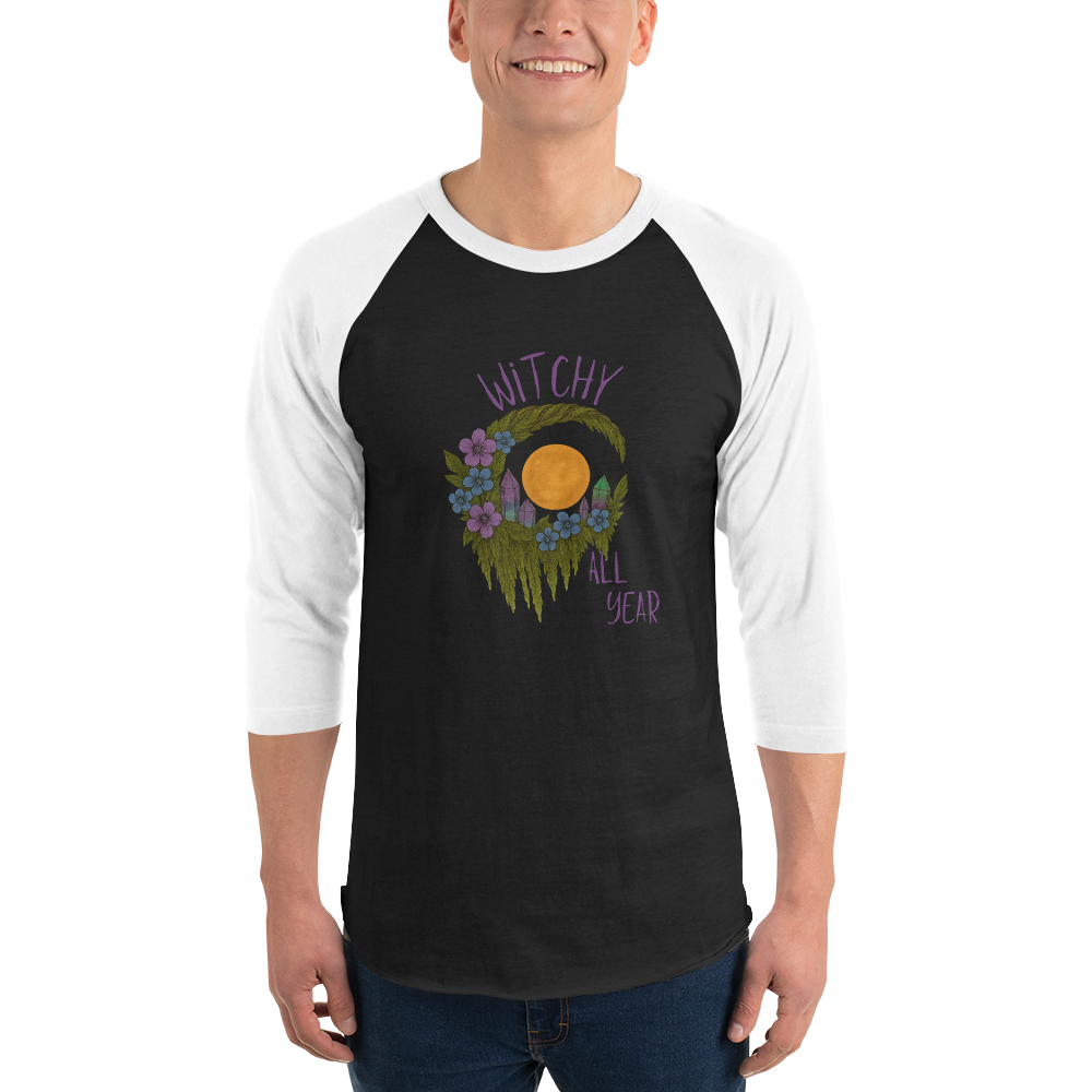 This is a man wearing a black and white raglan shirt with a design of a hand-drawn wreath made of moss-covered branches wraps around a glowing sun. The wreath is decorated with purple and blue flowers, small amethyst and fluorite crystals, and strands of hanging moss. The words “Witchy” curve above the wreath and “All Year” sits on the bottom right 