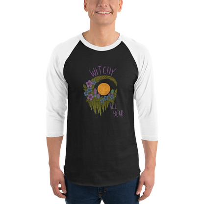 This is a man wearing a black and white raglan shirt with a design of a hand-drawn wreath made of moss-covered branches wraps around a glowing sun. The wreath is decorated with purple and blue flowers, small amethyst and fluorite crystals, and strands of hanging moss. The words “Witchy” curve above the wreath and “All Year” sits on the bottom right 