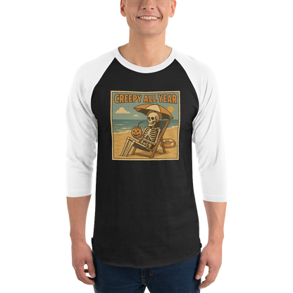 This is a man wearing a black and white raglan shirt with a retro-style illustration of a grinning skeleton lounging on a beach chair under an orange umbrella. The skeleton is holding a pumpkin-shaped drink cup with a straw and relaxing by the ocean, with a bright sun in the sky and a life preserver in the sand. The words “Creepy All Year” appear boldly at the top