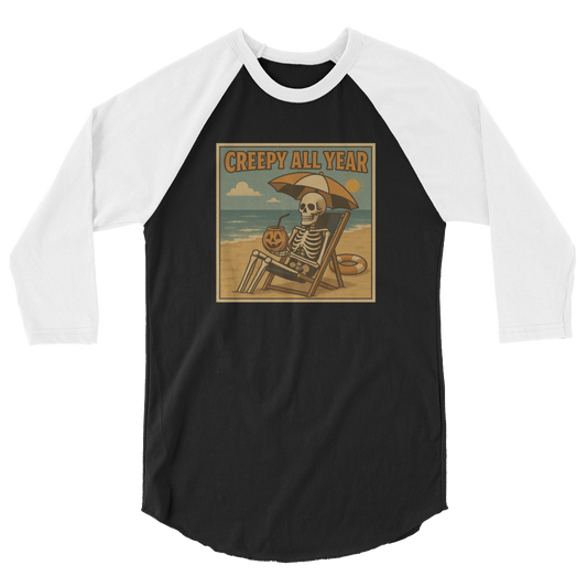 This is a black and white raglan shirt with a retro-style illustration of a grinning skeleton lounging on a beach chair under an orange umbrella. The skeleton is holding a pumpkin-shaped drink cup with a straw and relaxing by the ocean, with a bright sun in the sky and a life preserver in the sand. The words “Creepy All Year” appear boldly at the top