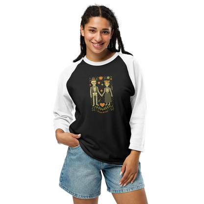 This is a woman with dark braids wearing a black and white raglan shirt with a Día de los Muertos–style illustration of a male and female skeleton holding hands, surrounded by marigolds, vines, and folk art patterns. The skeletons wear traditional black clothing, the man in a suit and hat, the woman with a floral crown with a red heart and leaves at the bottom and the text Creepy All Year. Rendered in warm tones of orange, green, and cream, symbolizing eternal love and remembrance