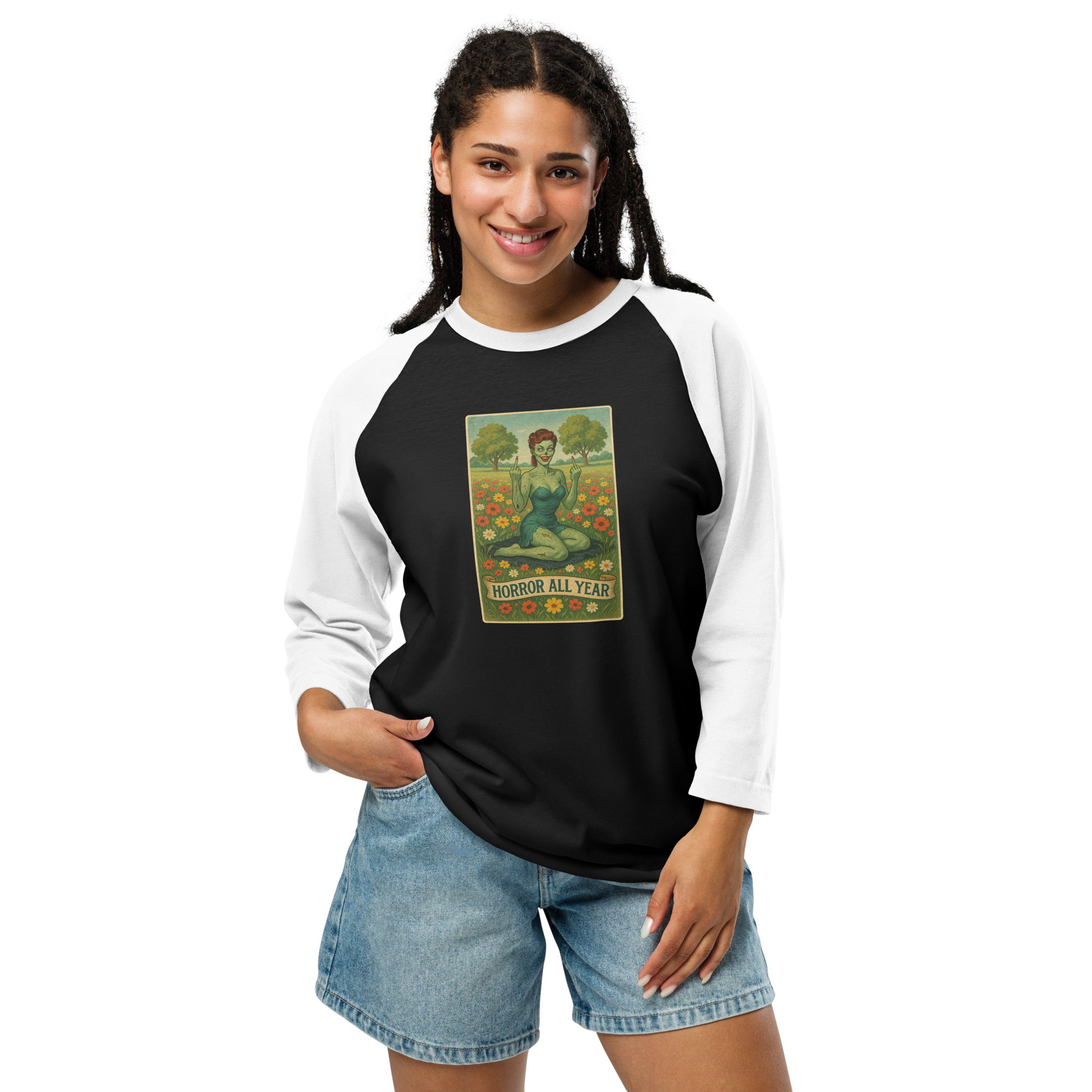 This is a woman with dark braids wearing a black and white raglan shirt with a design illustration of a sassy zombie pinup girl standing in a floral field, wearing tattered lingerie and flipping off the viewer with both hands. She has green skin, vintage curls, and a bold attitude. The design blends horror and humor, with a pastel color palette and creepy cute vibes