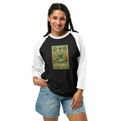 This is a woman with dark braids wearing a black and white raglan shirt with a design illustration of a sassy zombie pinup girl standing in a floral field, wearing tattered lingerie and flipping off the viewer with both hands. She has green skin, vintage curls, and a bold attitude. The design blends horror and humor, with a pastel color palette and creepy cute vibes