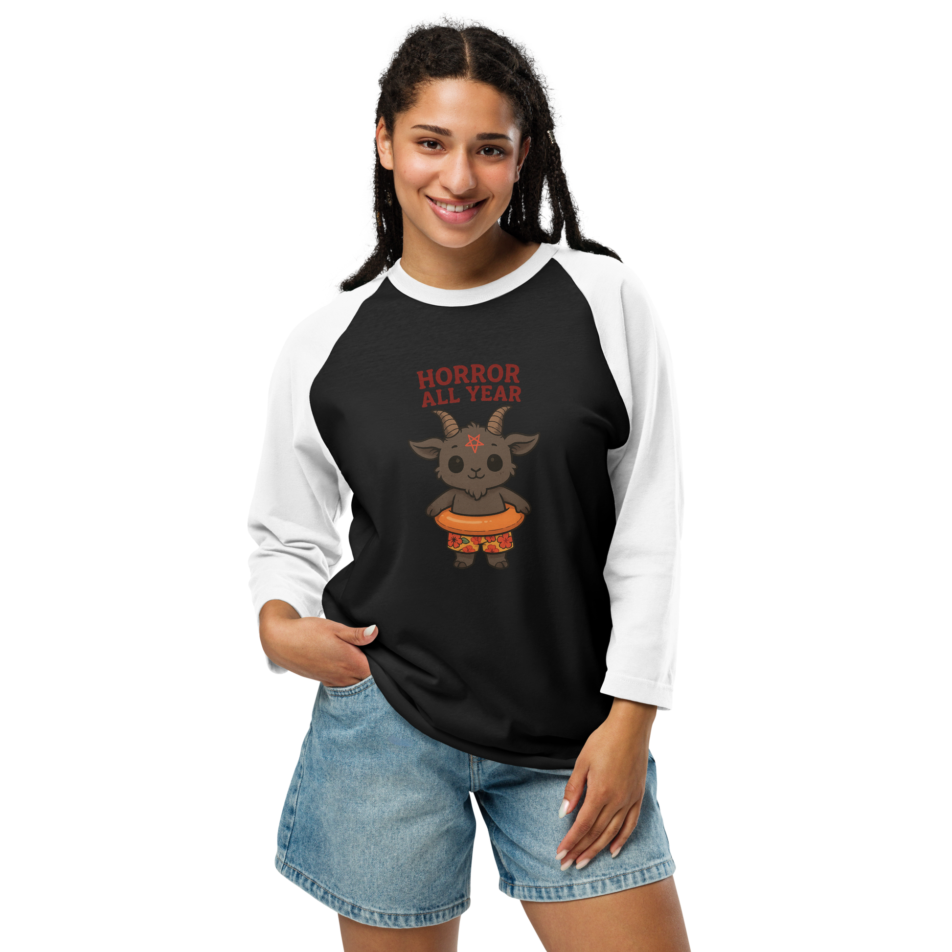 This is a woman with dark braids wearing a black and white raglan with a cartoon illustration of a baby Baphomet goat with curved horns and a playful grin, wearing bright floral swim trunks while floating in a colorful inner tube. The character is set against a light background with the bold text “Horror All Year” above in red letters
