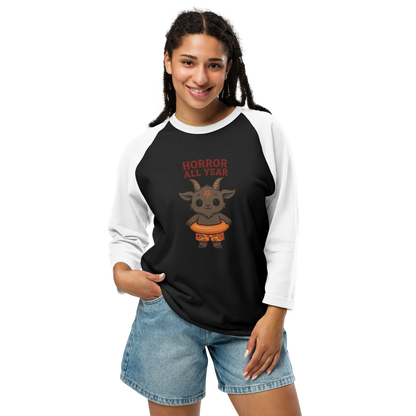 This is a woman with dark braids wearing a black and white raglan with a cartoon illustration of a baby Baphomet goat with curved horns and a playful grin, wearing bright floral swim trunks while floating in a colorful inner tube. The character is set against a light background with the bold text “Horror All Year” above in red letters