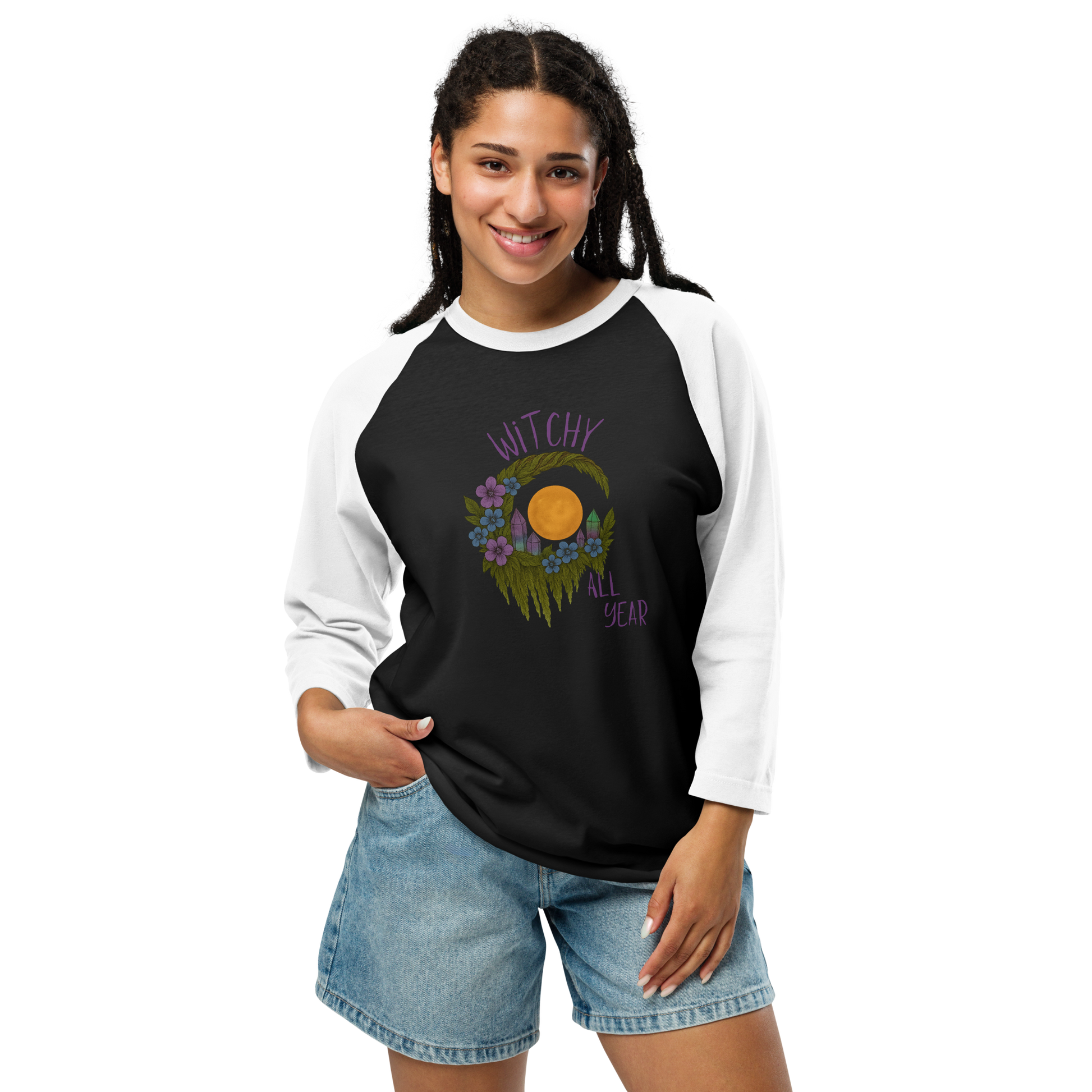 This is a woman with dark hair wearing a black and white raglan shirt with a design of a hand-drawn wreath made of moss-covered branches wraps around a glowing sun. The wreath is decorated with purple and blue flowers, small amethyst and fluorite crystals, and strands of hanging moss. The words “Witchy” curve above the wreath and “All Year” sits on the bottom right 