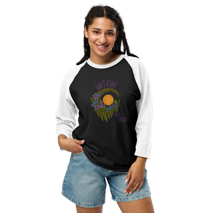 This is a woman with dark hair wearing a black and white raglan shirt with a design of a hand-drawn wreath made of moss-covered branches wraps around a glowing sun. The wreath is decorated with purple and blue flowers, small amethyst and fluorite crystals, and strands of hanging moss. The words “Witchy” curve above the wreath and “All Year” sits on the bottom right 