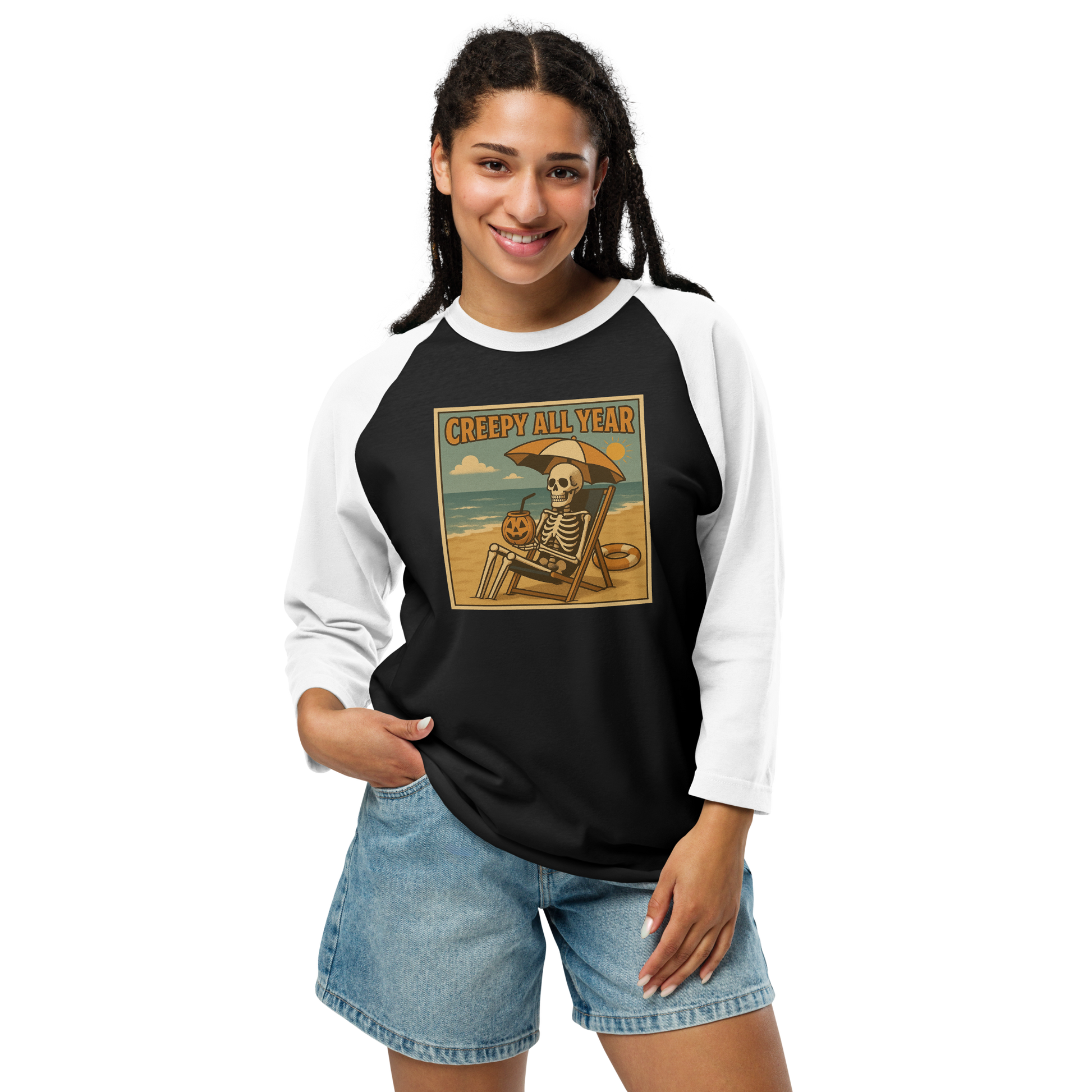This is a woman with dark hair wearing a black and white raglan shirt with a retro-style illustration of a grinning skeleton lounging on a beach chair under an orange umbrella. The skeleton is holding a pumpkin-shaped drink cup with a straw and relaxing by the ocean, with a bright sun in the sky and a life preserver in the sand. The words “Creepy All Year” appear boldly at the top