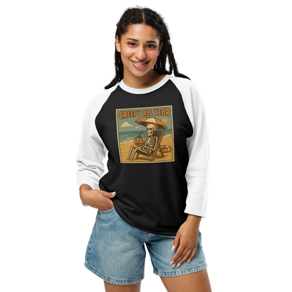 This is a woman with dark hair wearing a black and white raglan shirt with a retro-style illustration of a grinning skeleton lounging on a beach chair under an orange umbrella. The skeleton is holding a pumpkin-shaped drink cup with a straw and relaxing by the ocean, with a bright sun in the sky and a life preserver in the sand. The words “Creepy All Year” appear boldly at the top