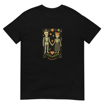 This is a black t-shirt with a Día de los Muertos–style illustration of a male and female skeleton holding hands, surrounded by marigolds, vines, and folk art patterns. The skeletons wear traditional black clothing, the man in a suit and hat, the woman with a floral crown with a red heart and leaves at the bottom and the text Creepy All Year. Rendered in warm tones of orange, green, and cream, symbolizing eternal love and remembrance