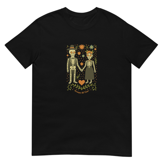 This is a black t-shirt with a Día de los Muertos–style illustration of a male and female skeleton holding hands, surrounded by marigolds, vines, and folk art patterns. The skeletons wear traditional black clothing, the man in a suit and hat, the woman with a floral crown with a red heart and leaves at the bottom and the text Creepy All Year. Rendered in warm tones of orange, green, and cream, symbolizing eternal love and remembrance