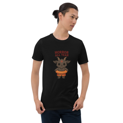 This is a man with dark hair wearing a black t-shirt with a cartoon illustration of a baby Baphomet goat with curved horns and a playful grin, wearing bright floral swim trunks while floating in a colorful inner tube. The character is set against a light background with the bold text “Horror All Year” above in red letters