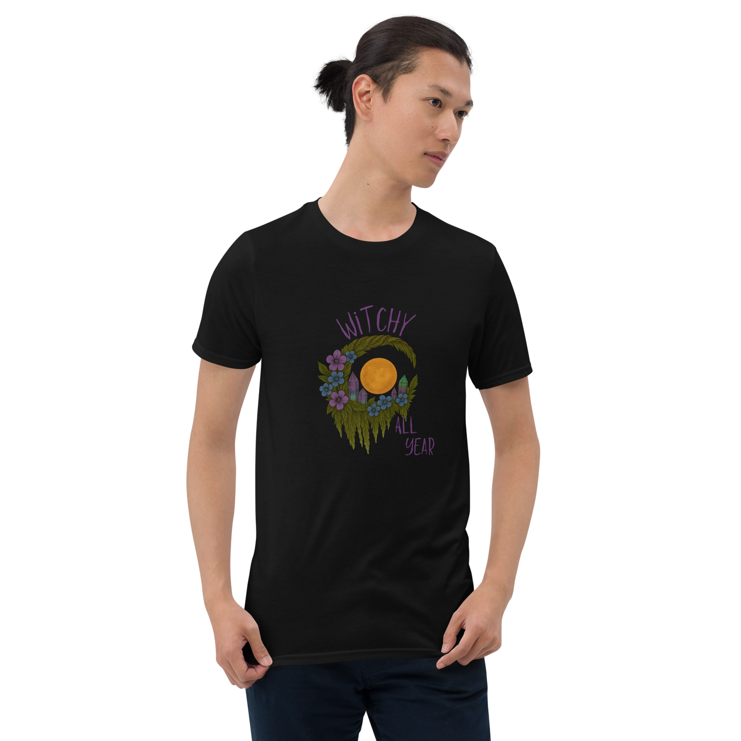 This is a man with dark hair wearing a black t-shirt with a design of a hand-drawn wreath made of moss-covered branches wraps around a glowing sun. The wreath is decorated with purple and blue flowers, small amethyst and fluorite crystals, and strands of hanging moss. The words “Witchy” curve above the wreath and “All Year” sits on the bottom right 