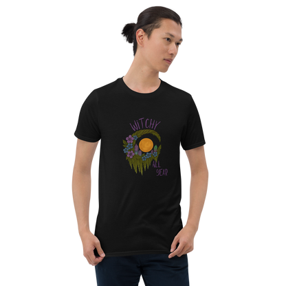 This is a man with dark hair wearing a black t-shirt with a design of a hand-drawn wreath made of moss-covered branches wraps around a glowing sun. The wreath is decorated with purple and blue flowers, small amethyst and fluorite crystals, and strands of hanging moss. The words “Witchy” curve above the wreath and “All Year” sits on the bottom right 