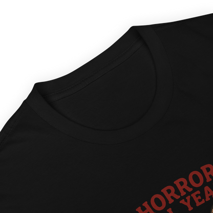 This is a zoom in of a black t-shirt with a cartoon illustration of a baby Baphomet goat with curved horns and a playful grin, wearing bright floral swim trunks while floating in a colorful inner tube. The character is set against a light background with the bold text “Horror All Year” above in red letters