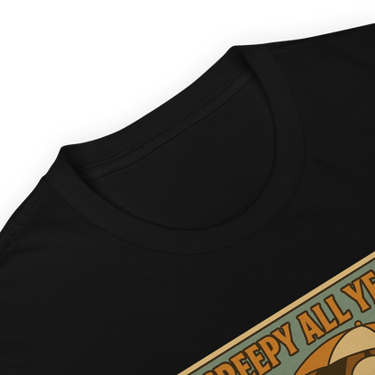 This is a zoom in of a black t-shirt with a retro-style illustration of a grinning skeleton lounging on a beach chair under an orange umbrella. The skeleton is holding a pumpkin-shaped drink cup with a straw and relaxing by the ocean, with a bright sun in the sky and a life preserver in the sand. The words “Creepy All Year” appear boldly at the top