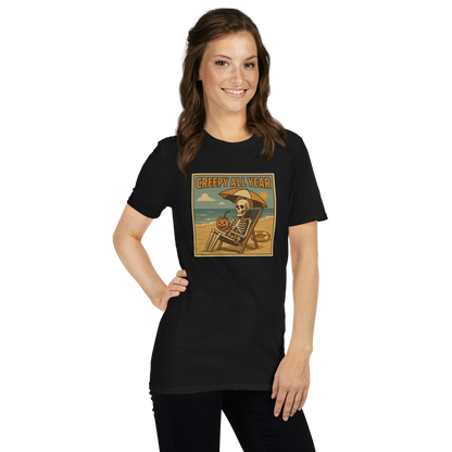 This is a woman with dark hair wearing a black t-shirt with a retro-style illustration of a grinning skeleton lounging on a beach chair under an orange umbrella. The skeleton is holding a pumpkin-shaped drink cup with a straw and relaxing by the ocean, with a bright sun in the sky and a life preserver in the sand. The words “Creepy All Year” appear boldly at the top