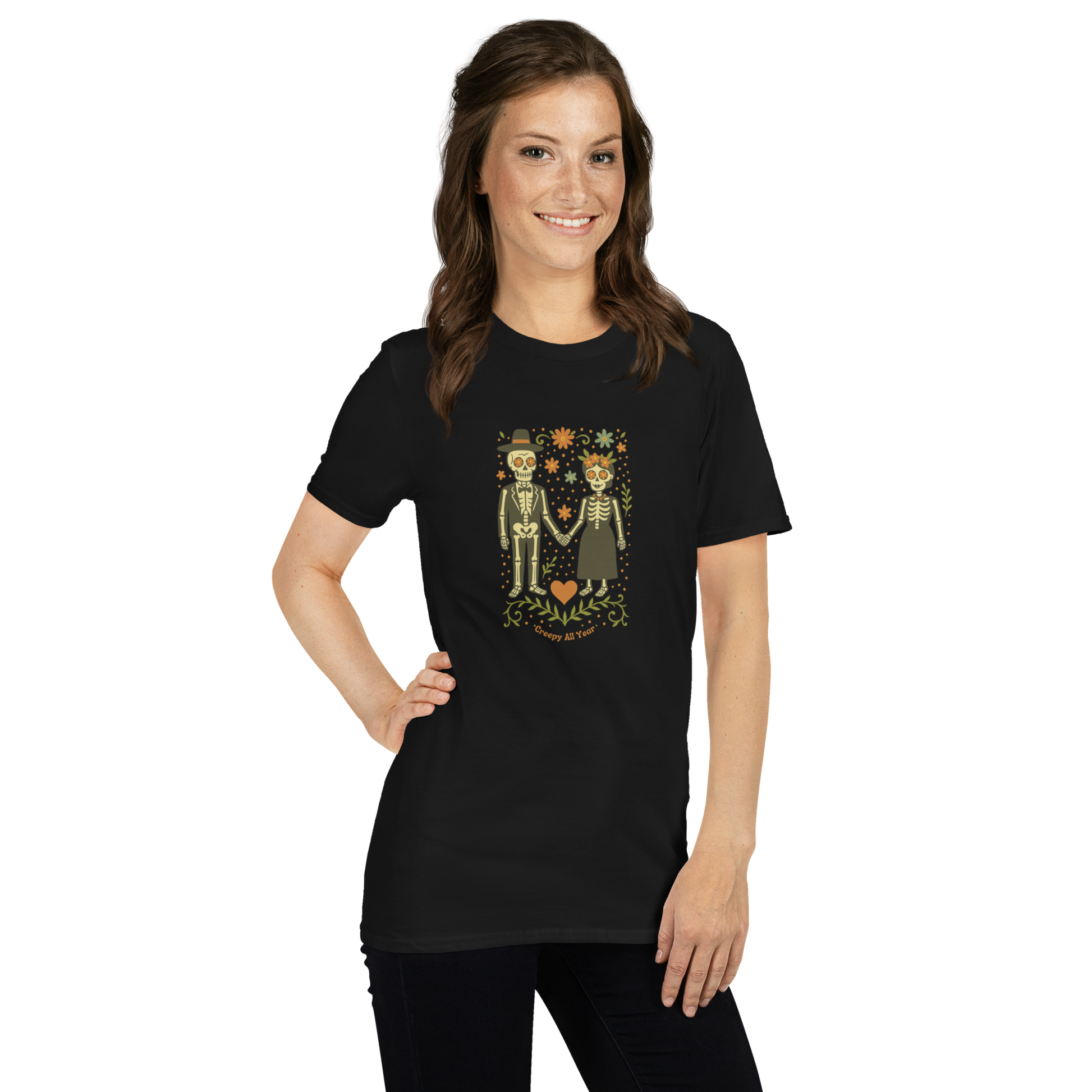 This is a woman with dark hair wearing a black t-shirt with a Día de los Muertos–style illustration of a male and female skeleton holding hands, surrounded by marigolds, vines, and folk art patterns. The skeletons wear traditional black clothing, the man in a suit and hat, the woman with a floral crown with a red heart and leaves at the bottom and the text Creepy All Year. Rendered in warm tones of orange, green, and cream, symbolizing eternal love and remembrance
