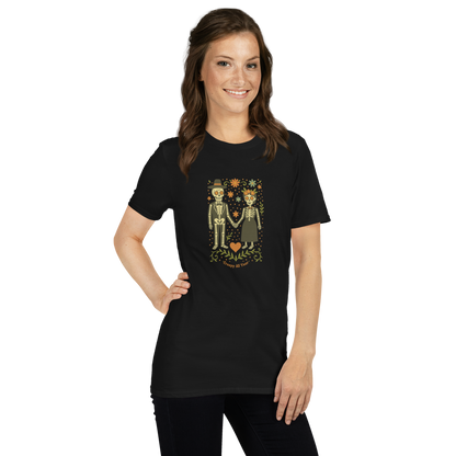 This is a woman with dark hair wearing a black t-shirt with a Día de los Muertos–style illustration of a male and female skeleton holding hands, surrounded by marigolds, vines, and folk art patterns. The skeletons wear traditional black clothing, the man in a suit and hat, the woman with a floral crown with a red heart and leaves at the bottom and the text Creepy All Year. Rendered in warm tones of orange, green, and cream, symbolizing eternal love and remembrance