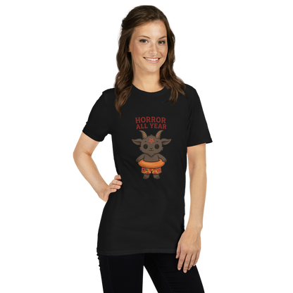 This is a woman with dark hair wearing a black t-shirt with a cartoon illustration of a baby Baphomet goat with curved horns and a playful grin, wearing bright floral swim trunks while floating in a colorful inner tube. The character is set against a light background with the bold text “Horror All Year” above in red letters