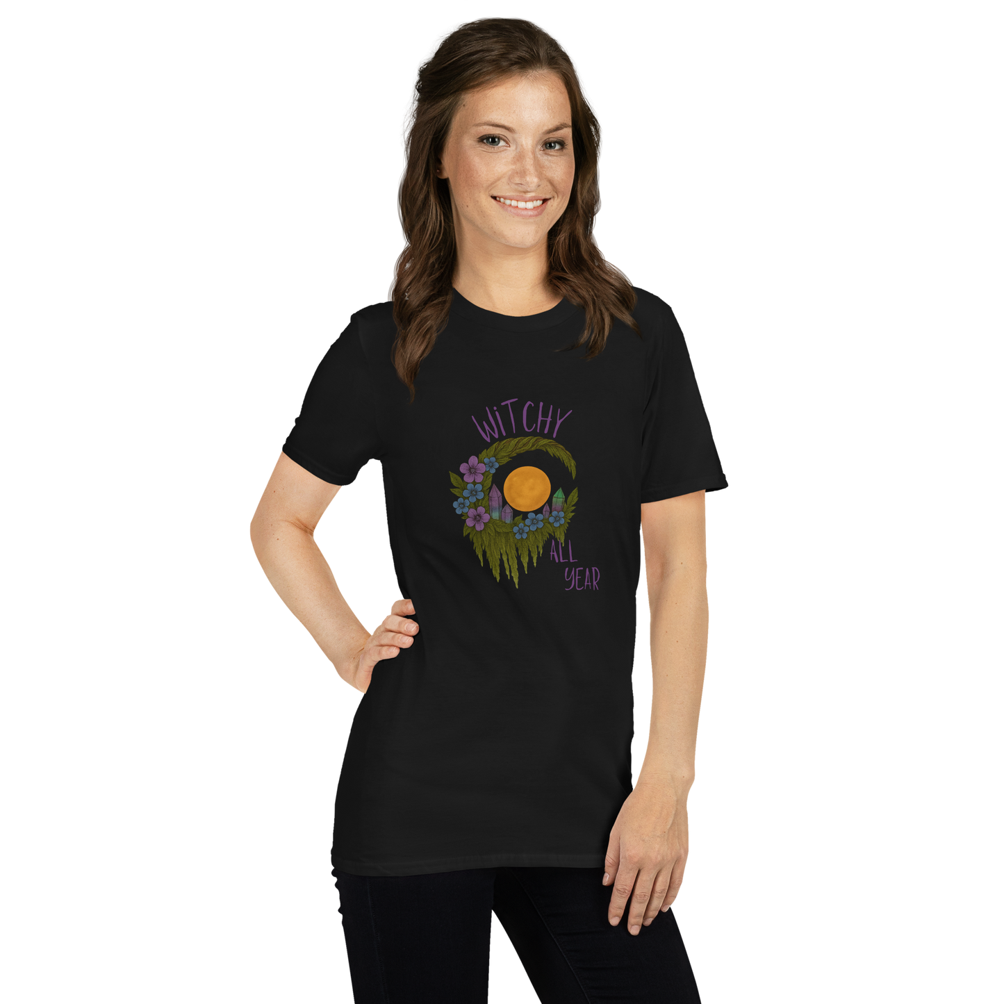 This is a woman with dark hair wearing a black t-shirt with a design of a hand-drawn wreath made of moss-covered branches wraps around a glowing sun. The wreath is decorated with purple and blue flowers, small amethyst and fluorite crystals, and strands of hanging moss. The words “Witchy” curve above the wreath and “All Year” sits on the bottom right 