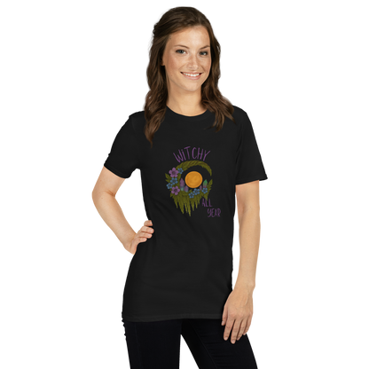 This is a woman with dark hair wearing a black t-shirt with a design of a hand-drawn wreath made of moss-covered branches wraps around a glowing sun. The wreath is decorated with purple and blue flowers, small amethyst and fluorite crystals, and strands of hanging moss. The words “Witchy” curve above the wreath and “All Year” sits on the bottom right 