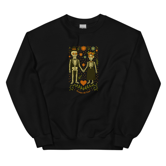 This is a black sweatshirt with a Día de los Muertos–style illustration of a male and female skeleton holding hands, surrounded by marigolds, vines, and folk art patterns. The skeletons wear traditional black clothing, the man in a suit and hat, the woman with a floral crown with a red heart and leaves at the bottom and the text Creepy All Year. Rendered in warm tones of orange, green, and cream, symbolizing eternal love and remembrance