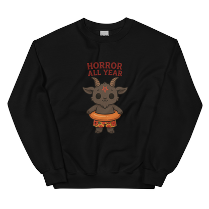 This is a black sweatshirt with a cartoon illustration of a baby Baphomet goat with curved horns and a playful grin, wearing bright floral swim trunks while floating in a colorful inner tube. The character is set against a light background with the bold text “Horror All Year” above in red letters
