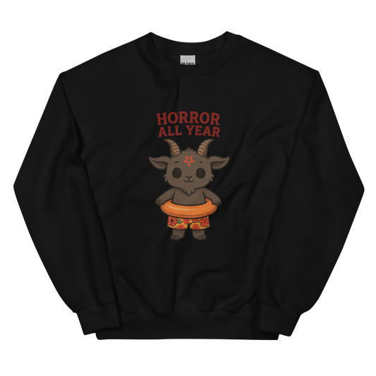 This is a black sweatshirt with a cartoon illustration of a baby Baphomet goat with curved horns and a playful grin, wearing bright floral swim trunks while floating in a colorful inner tube. The character is set against a light background with the bold text “Horror All Year” above in red letters
