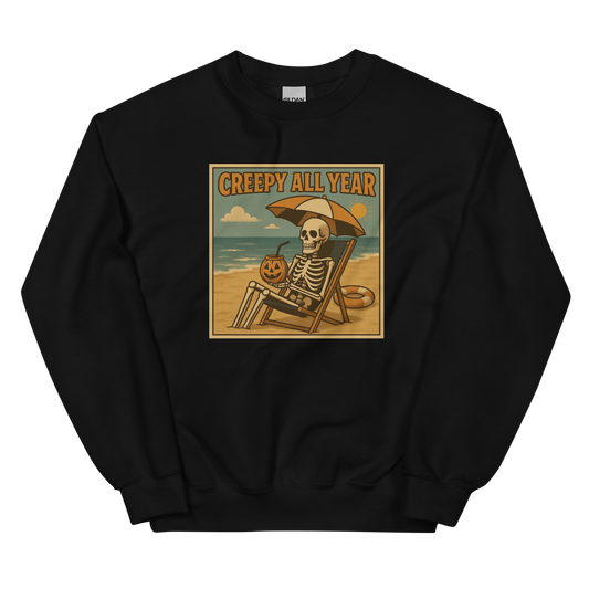 This is a black sweatshirt with a retro-style illustration of a grinning skeleton lounging on a beach chair under an orange umbrella. The skeleton is holding a pumpkin-shaped drink cup with a straw and relaxing by the ocean, with a bright sun in the sky and a life preserver in the sand. The words “Creepy All Year” appear boldly at the top