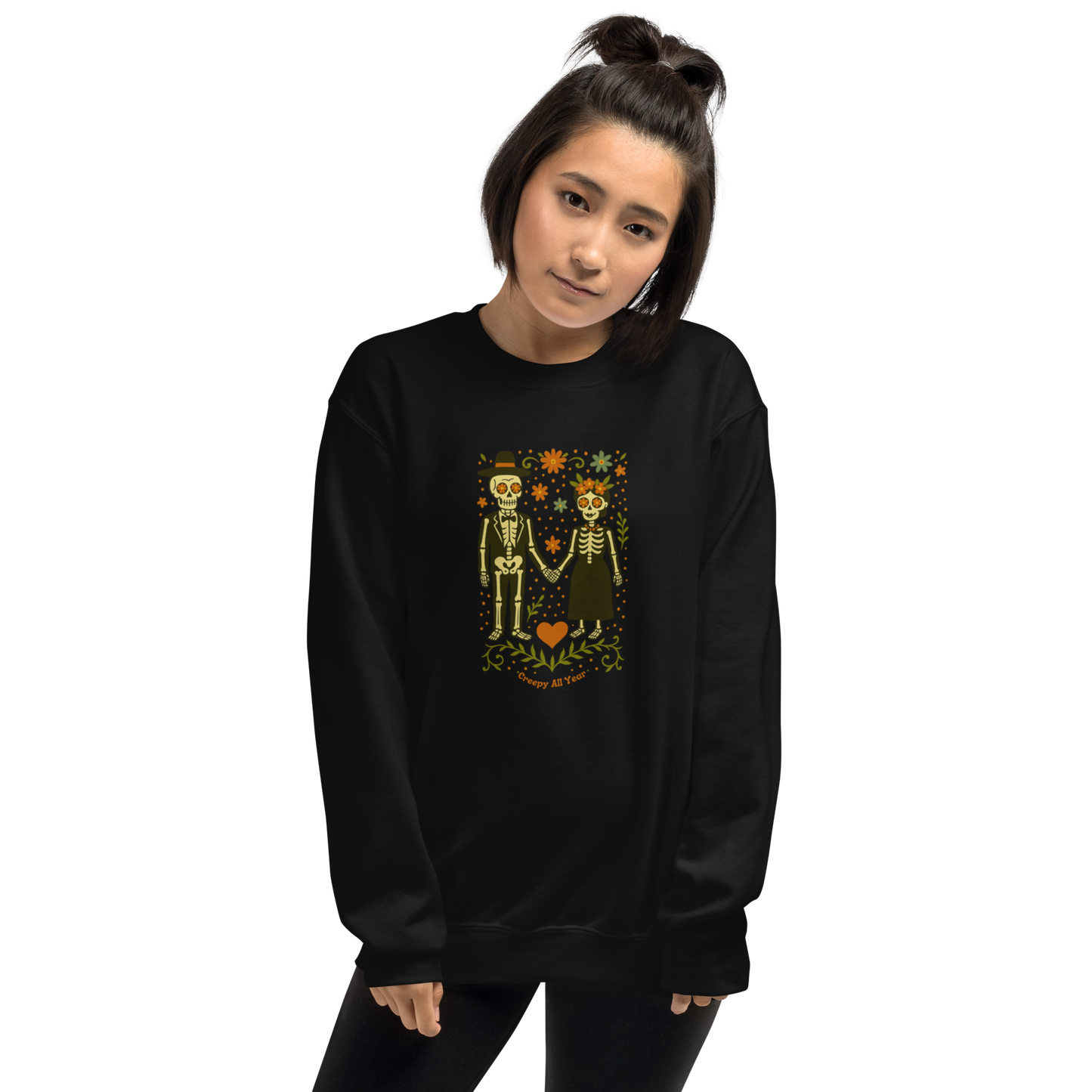 This is a woman with dark hair wearing a black sweatshirt with a Día de los Muertos–style illustration of a male and female skeleton holding hands, surrounded by marigolds, vines, and folk art patterns. The skeletons wear traditional black clothing, the man in a suit and hat, the woman with a floral crown with a red heart and leaves at the bottom and the text Creepy All Year. Rendered in warm tones of orange, green, and cream, symbolizing eternal love and remembrance