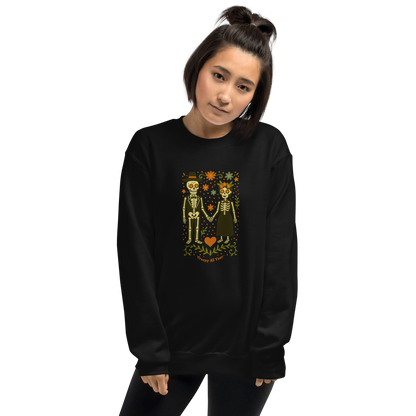 This is a woman with dark hair wearing a black sweatshirt with a Día de los Muertos–style illustration of a male and female skeleton holding hands, surrounded by marigolds, vines, and folk art patterns. The skeletons wear traditional black clothing, the man in a suit and hat, the woman with a floral crown with a red heart and leaves at the bottom and the text Creepy All Year. Rendered in warm tones of orange, green, and cream, symbolizing eternal love and remembrance