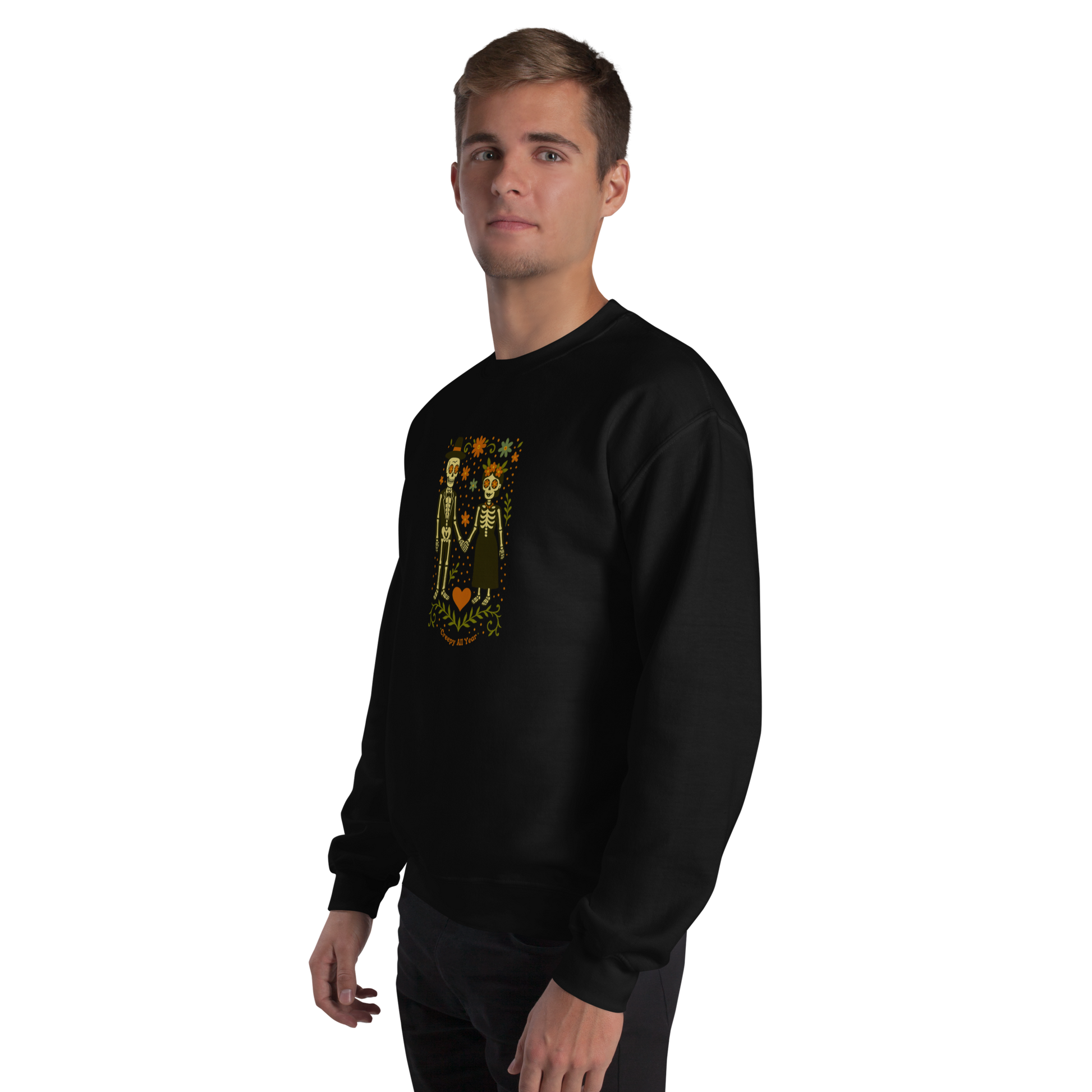 This is a man with blonde hair wearing a black sweatshirt with a Día de los Muertos–style illustration of a male and female skeleton holding hands, surrounded by marigolds, vines, and folk art patterns. The skeletons wear traditional black clothing, the man in a suit and hat, the woman with a floral crown with a red heart and leaves at the bottom and the text Creepy All Year. Rendered in warm tones of orange, green, and cream, symbolizing eternal love and remembrance