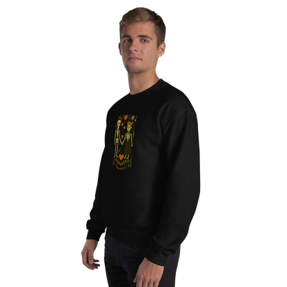 This is a man with blonde hair wearing a black sweatshirt with a Día de los Muertos–style illustration of a male and female skeleton holding hands, surrounded by marigolds, vines, and folk art patterns. The skeletons wear traditional black clothing, the man in a suit and hat, the woman with a floral crown with a red heart and leaves at the bottom and the text Creepy All Year. Rendered in warm tones of orange, green, and cream, symbolizing eternal love and remembrance