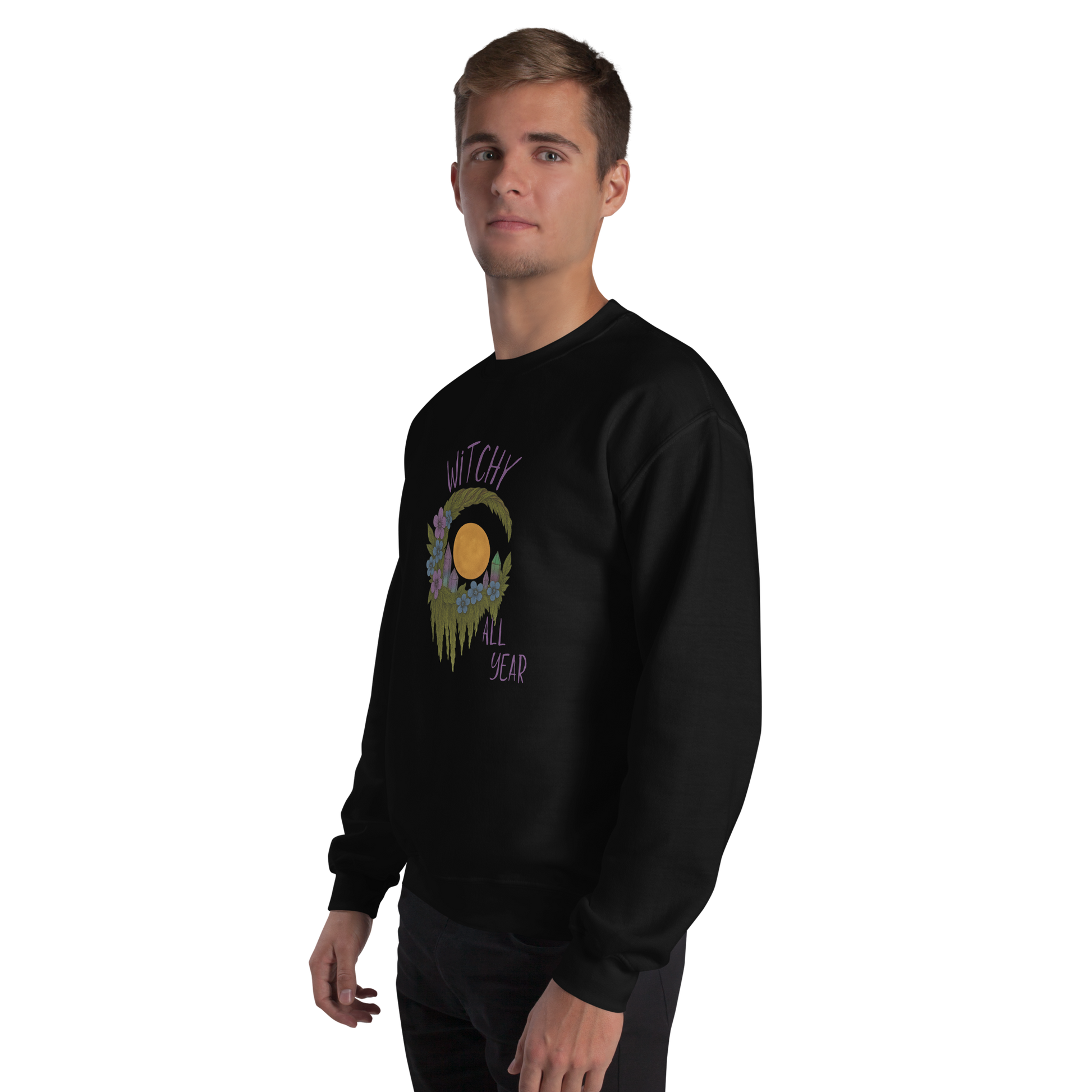 This is a man with blonde hair wearing a black sweatshirt with a design of a hand-drawn wreath made of moss-covered branches wraps around a glowing sun. The wreath is decorated with purple and blue flowers, small amethyst and fluorite crystals, and strands of hanging moss. The words “Witchy” curve above the wreath and “All Year” sits on the bottom right 