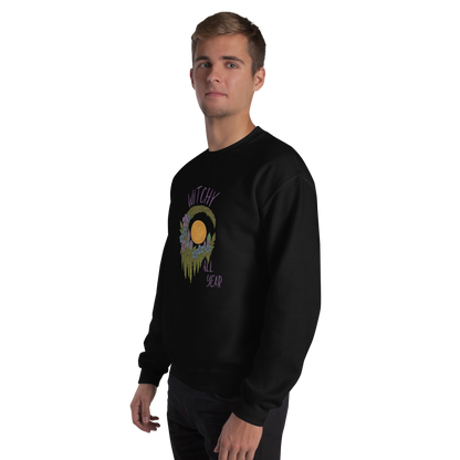 This is a man with blonde hair wearing a black sweatshirt with a design of a hand-drawn wreath made of moss-covered branches wraps around a glowing sun. The wreath is decorated with purple and blue flowers, small amethyst and fluorite crystals, and strands of hanging moss. The words “Witchy” curve above the wreath and “All Year” sits on the bottom right 