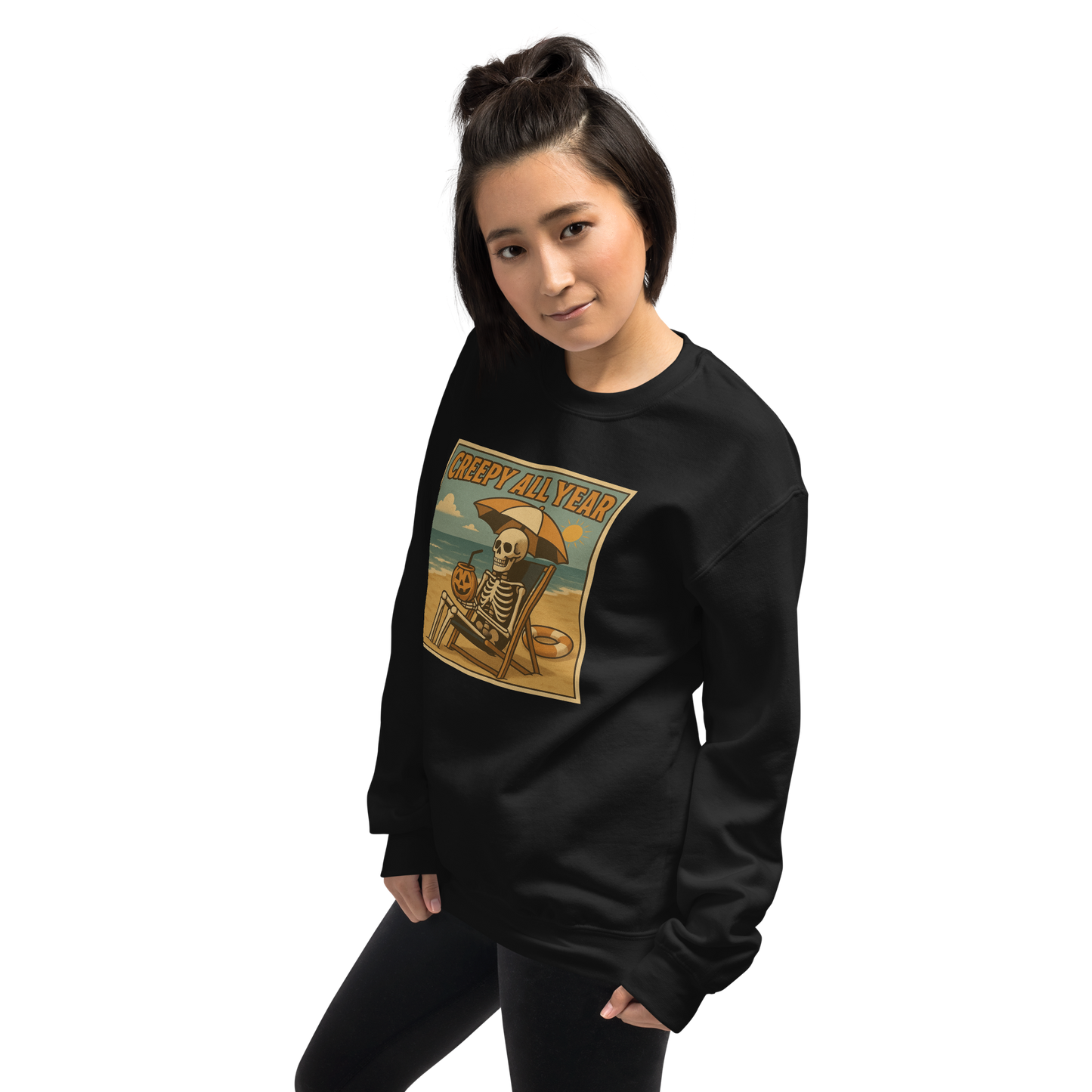 This is a woman with dark hair wearing a black sweatshirt with a retro-style illustration of a grinning skeleton lounging on a beach chair under an orange umbrella. The skeleton is holding a pumpkin-shaped drink cup with a straw and relaxing by the ocean, with a bright sun in the sky and a life preserver in the sand. The words “Creepy All Year” appear boldly at the top