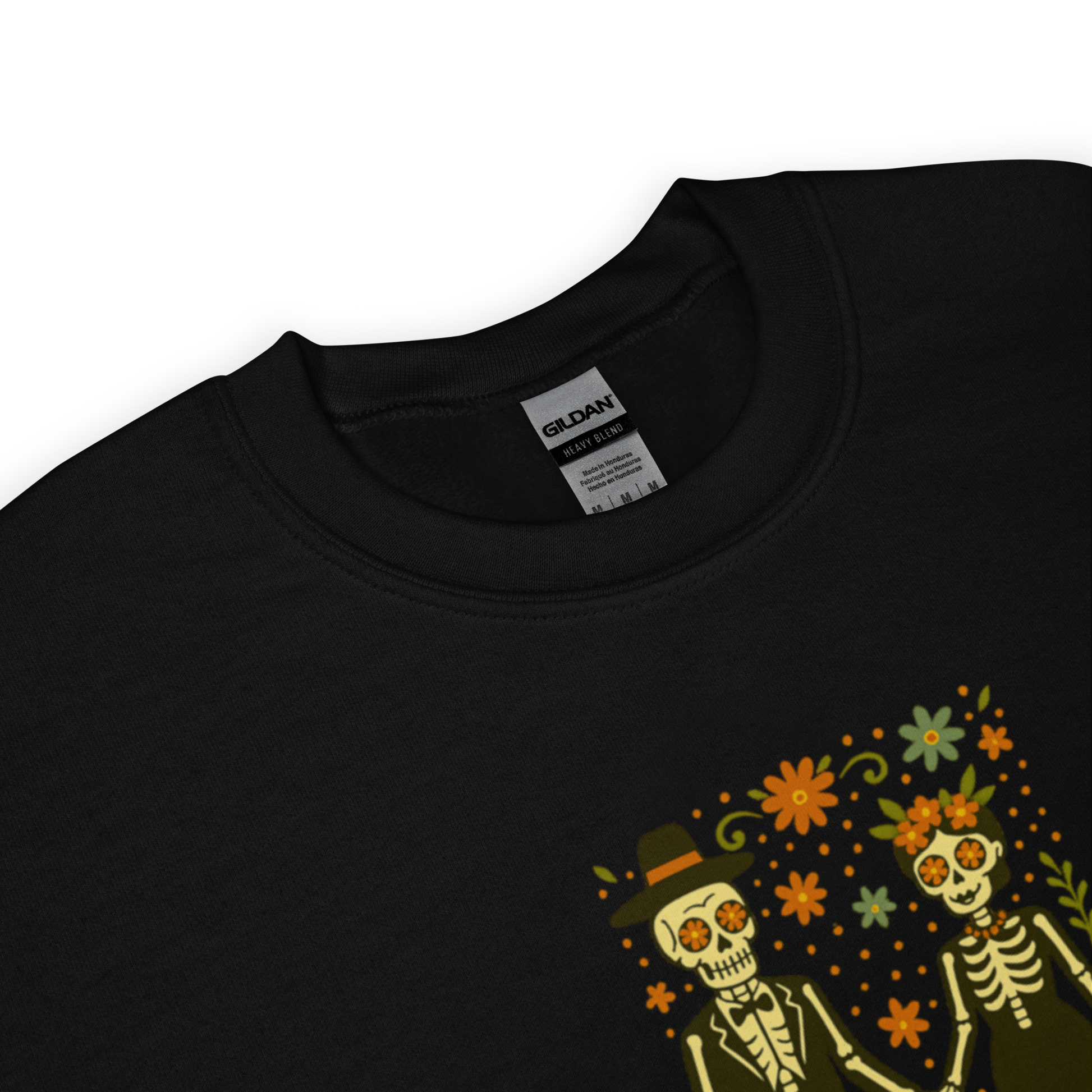 This is a zoom in on a black sweatshirt with a Día de los Muertos–style illustration of a male and female skeleton holding hands, surrounded by marigolds, vines, and folk art patterns. The skeletons wear traditional black clothing, the man in a suit and hat, the woman with a floral crown with a red heart and leaves at the bottom and the text Creepy All Year. Rendered in warm tones of orange, green, and cream, symbolizing eternal love and remembrance